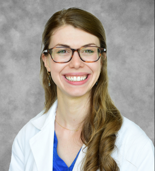 A white woman with long brown hair parted on the left, hanging down her right shoulder, wearing large frame brown glasses and a blue shirt under a white doctor's coat