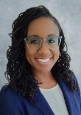 A Black woman with long black hair in microlocs parted on the right, wearing large clear framed glasses and a white shirt under a blue blazer