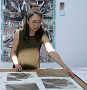 A Korean woman with dark brown hair half up in a bun, wearing glasses and a sleeveless green brown dress, leaning over a table with several photo prints on it