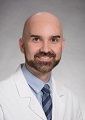 A smiling, bald, white man with a neatly trimmed brown beard and mustache wearing a light blue button down shirt and white doctor's coat