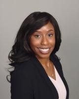 A smiling Black woman with slightly curled black hair parted to the left wearing a black blazer with white V neck shirt